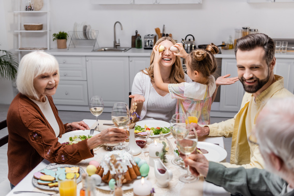 girl covering eyes of mother with easter eggs while family toasting with wine glasses - Fotó, kép