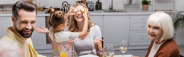 chica cubriendo los ojos de feliz mamá mientras se divierten durante la cena de Pascua con la familia, pancarta - Foto, Imagen