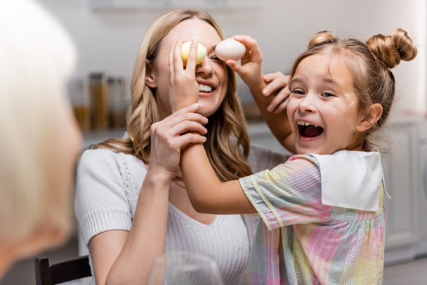 excitada chica cubriendo los ojos de la madre con huevos de Pascua cerca borrosa abuelita - Foto, Imagen