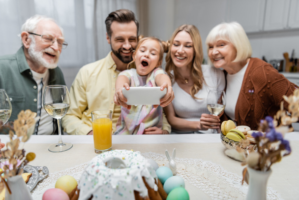 excited girl with open mouth taking selfie on smartphone during easter dinner with family - Foto, Imagen