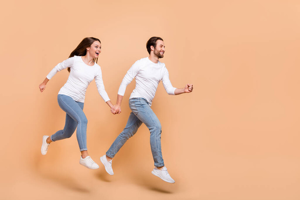 Full length body size view of attractive cheerful partners jumping holding hands going isolated over beige pastel color background - Foto, afbeelding