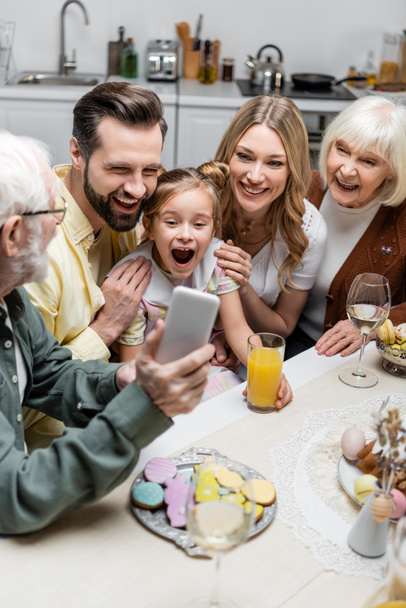 senior man holding smartphone near excited girl with open mouth and happy family - Foto, Imagen