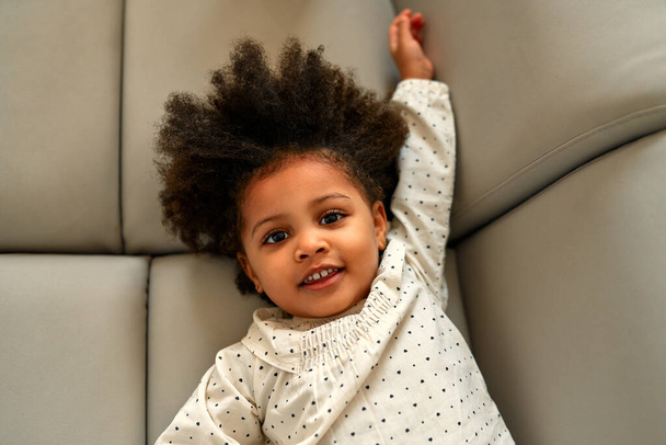 Słodkie afrykańskie dziecko dziewczyna z kręcone puszyste włosy leżące na kanapie w sukience w salonie w domu. - Zdjęcie, obraz
