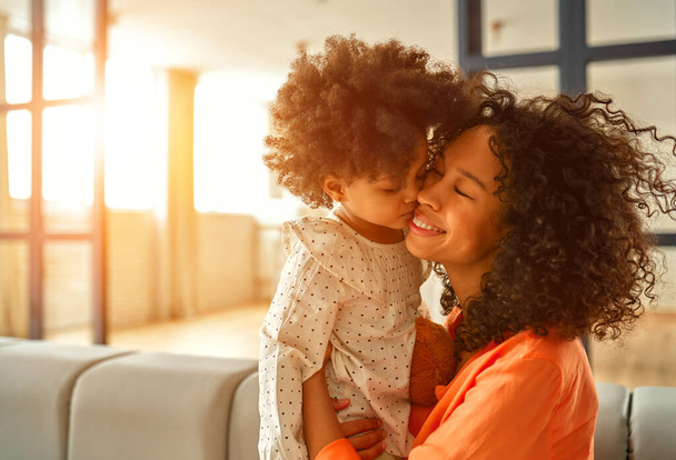 アフリカ系アメリカ人女性とともに彼女の小さな娘とともに巻き毛座っていますソファと優しく抱擁で自宅のリビングルーム. - 写真・画像