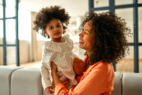 Afro-Amerikaanse vrouw met haar dochtertje met krullend pluizig haar zitten op de bank en plezier hebben in de woonkamer thuis. - Foto, afbeelding