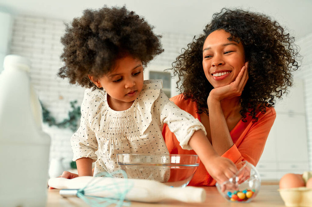 Afro-Amerikaanse vrouw met haar dochtertje met krullend pluizig haar die plezier heeft en gebak maakt in de keuken. Moeder en dochter koken samen. - Foto, afbeelding
