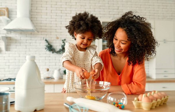 Afrikai amerikai nő a kislányával, göndör, bolyhos hajjal, jól szórakozik és süteményeket készít a konyhában. Anya és lánya együtt főznek.. - Fotó, kép