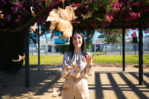 Όμορφη μελαχρινή κοπέλα με ανοιχτές αγκάλες και πεσμένα φύλλα στο πάρκο το φθινόπωρο. Μια γυναίκα πετάει φύλλα. Φθινοπωρινή ιδέα - Φωτογραφία, εικόνα