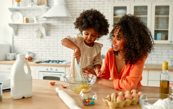 Kıvırcık saçlı ve mutfakta hamur işi yapan küçük kızı olan Afro-Amerikalı bir kadın. Anne ve kızı birlikte yemek pişiriyor.. - Fotoğraf, Görsel