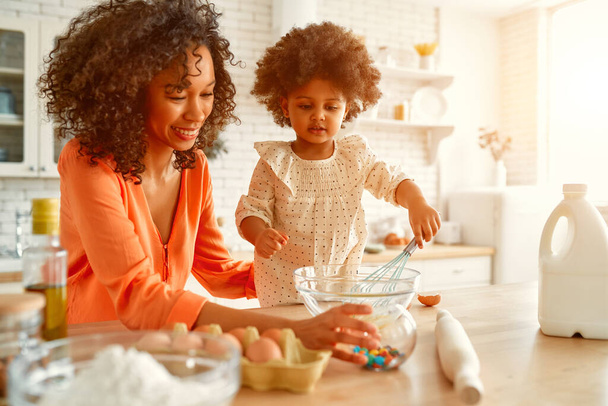 African American woman with her little daughter with curly fluffy hair having fun and making pastries in the kitchen. Mom and daughter cooking together. - Photo, Image