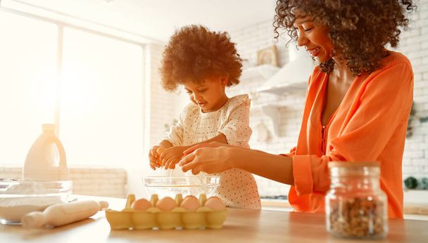 Afroamerikanerin mit ihrer kleinen Tochter mit lockigem, flauschigem Haar, die Spaß hat und in der Küche Gebäck backt. Mutter und Tochter kochen zusammen. - Foto, Bild