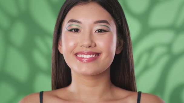 Молодий і красивий. Молода щаслива азіатська жінка з креативним макіяжем посміхається до камери, зелений фон, повільний рух
 - Кадри, відео