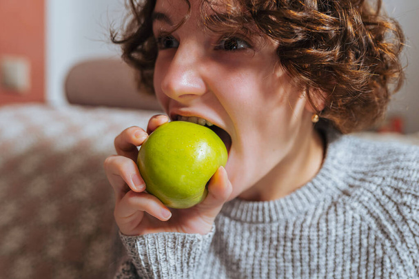 Kahverengi kıvırcık saçlı genç kız evde otururken yeşil elma yiyor. Yeşil elmayı iştahla yiyen genç kız duygularını ifade ediyor. - Fotoğraf, Görsel