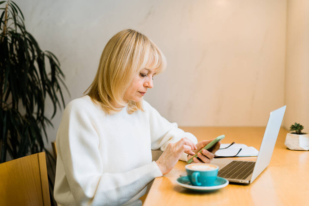 Kypsä aikuinen nainen istuu kahvilassa kahvimukin kanssa ja työskentelee verkossa kannettavalla tietokoneella. Liikenainen kutoma viestejä matkapuhelimeen coworking tilaa paahtimet kahvila - Valokuva, kuva