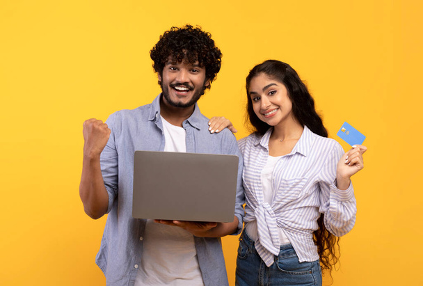 Ja, riesige Umsätze im Webstore. Glückliches indisches Paar kauft online ein, hält Kreditkarte und benutzt Laptop - Foto, Bild