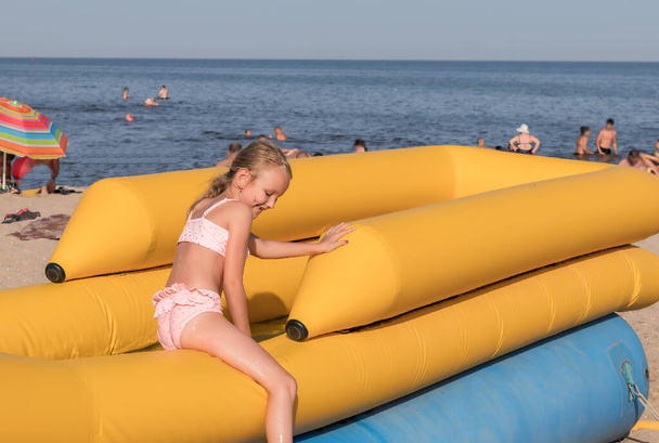 6-7 yaşlarında bir kız, sarışın, kumsalda oynuyor, şişme slaytlara biniyor, uzaklardan denizi görebilirsiniz. - Fotoğraf, Görsel