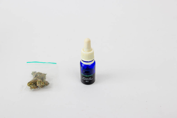 Gros plan d'un gramme de bourgeons secs de marijuana médicale dans un sac en plastique transparent, bouteille en verre bleu avec 10 ml d'huile de CBD, extrait de cannabis sur fond de studio blanc. Concept de phytothérapie alternative - Photo, image