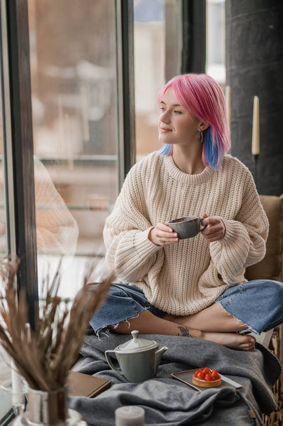 Молодая девушка с розовыми волосами пьет чай у окна - Фото, изображение