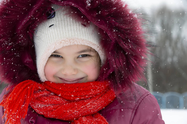 Dziecko w zimie. Mała dziewczynka w ciepłym kapeluszu i kapturze patrzy w kamerę i uśmiecha się. - Zdjęcie, obraz