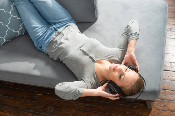 Young beautiful girl lies on a sofa in headphones and listens to music. Happy blond woman is resting at home. - Foto, imagen
