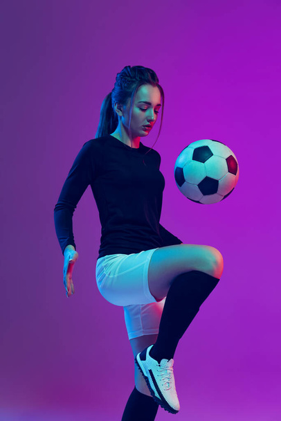 Emotive girl, female soccer, football player in motion, action isolated on purple studio background in neon light. Sport, active, healthy lifestyle - Zdjęcie, obraz