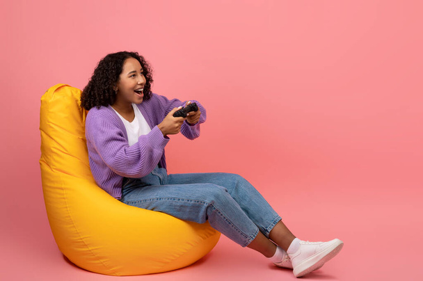 Excited black female gamer with joystick playing online computer game, sitting in bean bag chair on pink background - Photo, Image