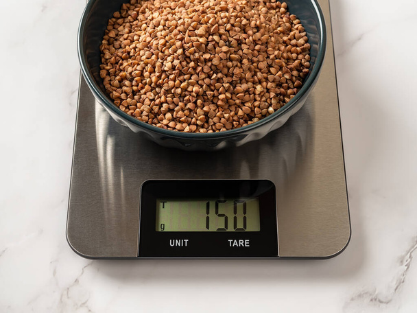 Raw organic buckwheat groats in a bowl on a digital kitchen scale. 150g roasted wholegrain buckwheat seeds on the scales displaying grams. Gluten free diet and dietary fiber concepts. Top view. - Valokuva, kuva