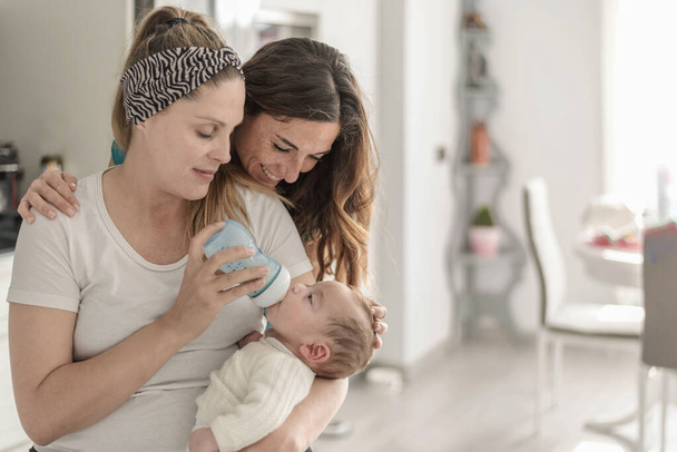 Um casal de lésbicas dando uma garrafa de leite para seu bebê, casamento gay, pais gays e conceito de adoção - Foto, Imagem