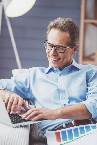 Handsome middle aged businessman in classic shirt and eyeglasses is using a laptop and smiling while working at home - Zdjęcie, obraz