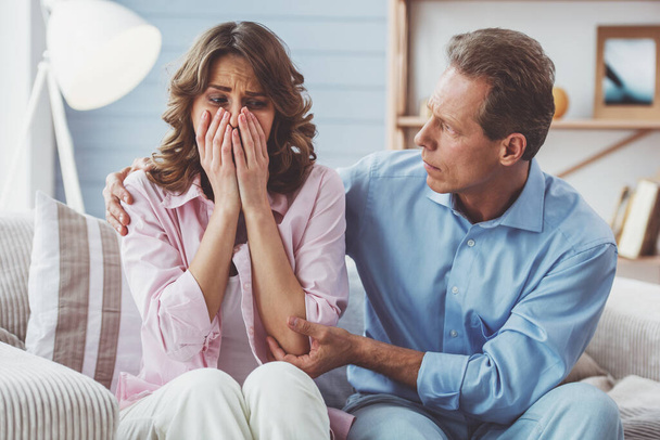 Mujer atractiva está llorando mientras su hombre la calma, sentado en el sofá en casa
 - Foto, imagen