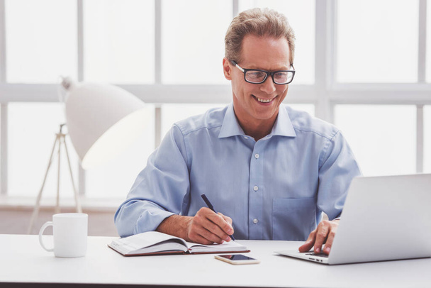 Handsome middle aged businessman in classic shirt and eyeglasses is using a laptop, taking notes and smiling while working in office - Fotó, kép