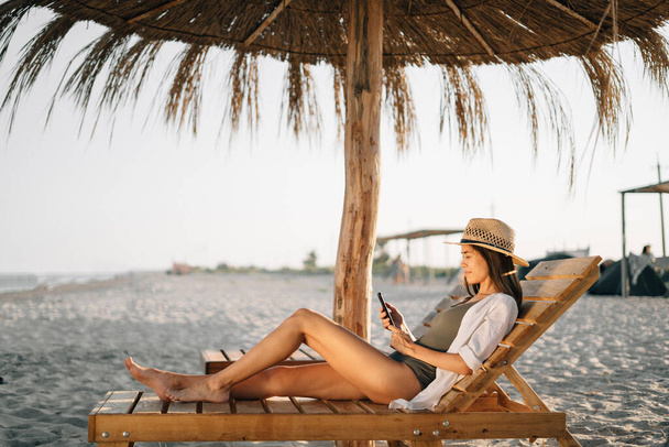 Vacaciones por mar, fin de semana en la playa, una hermosa mujer bronceada en un sombrero mira a la pantalla del teléfono inteligente, se sienta en una tumbona en la playa bajo un paraguas de paja. Concepto de vacaciones. espacio de copia. - Foto, Imagen