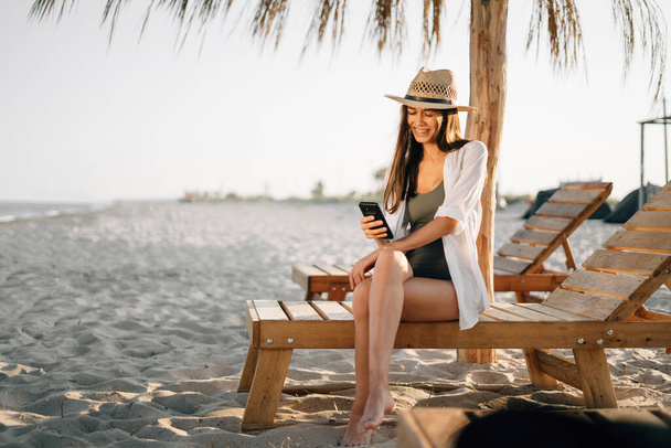 Ein junges Mädchen im Tropenresort sitzt während ihres Urlaubs auf einem hölzernen Liegestuhl in der Nähe von Strohschirmen am Strand, hält ein Smartphone in der Hand und macht mit der Anwendung einen Videoanruf. Kopierraum - Foto, Bild