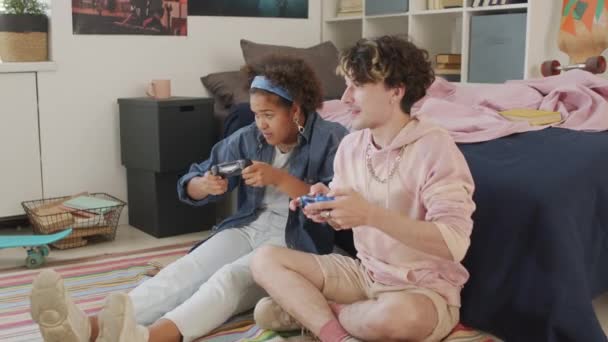Langzame beweging van blanke mannelijke en zwarte vrouwelijke klasgenoten zittend op de slaapkamervloer overdag, met behulp van game console controllers, spelen, en plezier hebben - Video