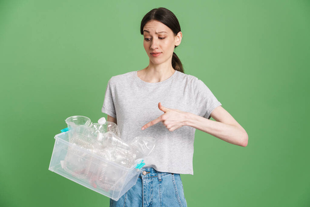 Young brunette woman holding and pointing finger at plastic waste isolated over green background - Foto, Imagem