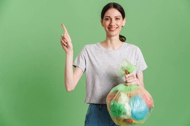 Young woman pointing finger upward while posing with plastic bags isolated over green background - Foto, afbeelding