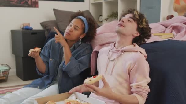Medium lang van zwart meisje en haar mannelijke blanke vriend zitten op de slaapkamer vloer overdag, chatten, glimlachen en eten pizza uit doos, met een snack na school - Video