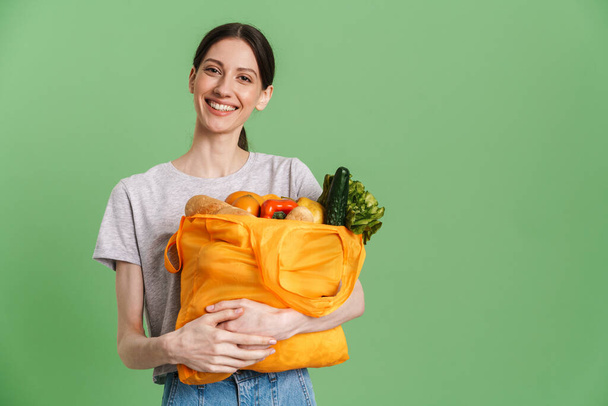 Young brunette woman smiling while holding textile bag with vegetable isolated over green background - Foto, imagen