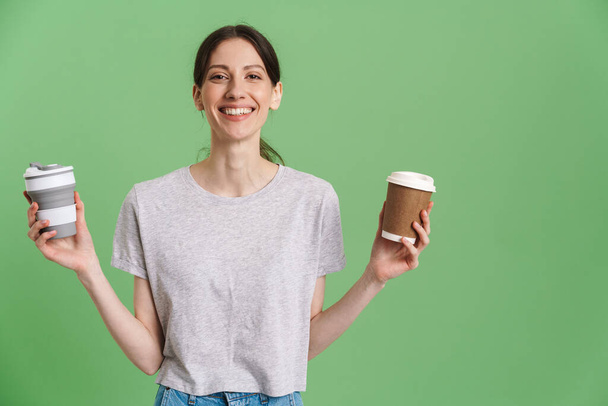 Young brunette woman smiling while posing with takeaway cups isolated over green background - Foto, afbeelding