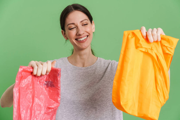 Young brunette woman showing plastic and textile bags isolated over green background - Foto, imagen