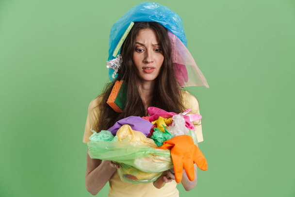 Young brunette woman with plastic waste expressing surprise at camera isolated over green background - Photo, image