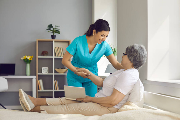 Happy elderly woman patient lying on bed with book talking to caregiver - Photo, Image