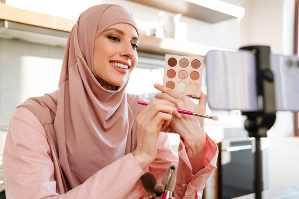 Young muslim woman in hijab vlogging herself about beauty products with making a video for her blog on cosmetics using phone camera at home - Zdjęcie, obraz
