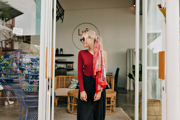 Pretty young woman with blond hair wearing cherry sweater and black trousers looking aside and smiling on background of modern stylish cafeteria. High quality photo - Fotoğraf, Görsel