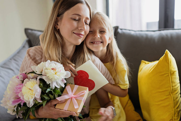 Child daughter congratulates mother and gives present card, gift and bouquet of flowers at home. Mom and girl smiling and hugging on couch. Happy mothers day celebration - Fotografie, Obrázek