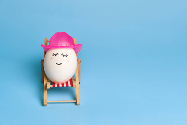 Creative funny composition with Easter egg wearing a hat sitting on deck chair on bright blue background. Trendy minimal spring or summer vacation, holiday and travel concept. Copy space - Foto, imagen