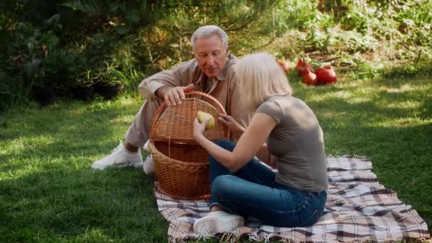 Szczęśliwa para seniorów o piknik na świeżym powietrzu, relaks na kocu w ogrodzie, wyjmowanie i wąchanie świeżych owoców z kosza, wspólne spędzanie czasu na łonie natury, powolny film wideo - Materiał filmowy, wideo