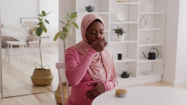 Szczęśliwy w ciąży czarny kobieta jedzenie chia budyń noszenie hidżab w pomieszczeniach - Materiał filmowy, wideo