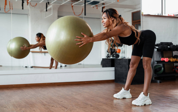 Athletic physique, cardio training for burning calories with a ball. A healthy lifestyle at a young age. A woman does a body stretching workout in a bright fitness studio. - Foto, imagen