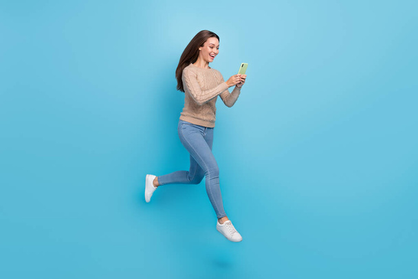 Full length body size view of attractive cheery focused girl jumping using gadget isolated over bright blue color background - 写真・画像